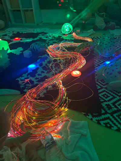 This media file depicts a sensory play setup, likely in a home or therapeutic environment in Wakefield, United Kingdom. The primary subjects are a large bundle of colorful fiber optic strands, illuminated by various colored lights. Red, orange, yellow, and blue lights are visible, casting a vibrant and dynamic glow. Scattered among the fiber optics are several objects, including inflatable balloons with patterns, a white blanket, and what appears to be a partially filled plastic water bottle. A child's hand is visible in the lower right corner, interacting with the fiber optic strands, suggesting an activity of exploration or play. The overall scene is dimly lit, emphasizing the illuminated elements and creating a cozy, engaging atmosphere. The background includes patterned fabric and shelving units, further reinforcing the idea of a dedicated play or sensory space. The emotion conveyed is one of wonder and sensory engagement, typical of a child interacting with a stimulating environment. There is no discernible time of day or weather information, but the indoor setting is evident. Visible text on the water bottle includes "PENNY'S".
