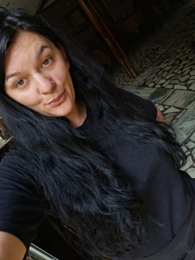 A woman with long, dark hair is taking a selfie. She is wearing a black t-shirt and has light brown eyes and a light complexion. Her head is tilted slightly and she is looking towards the camera with a slight smile. Her hair is flowing around her face and shoulders. The background appears to be indoors, with a tiled floor and hints of furniture and a window. The lighting suggests it could be daytime. The overall scene is casual and personal.