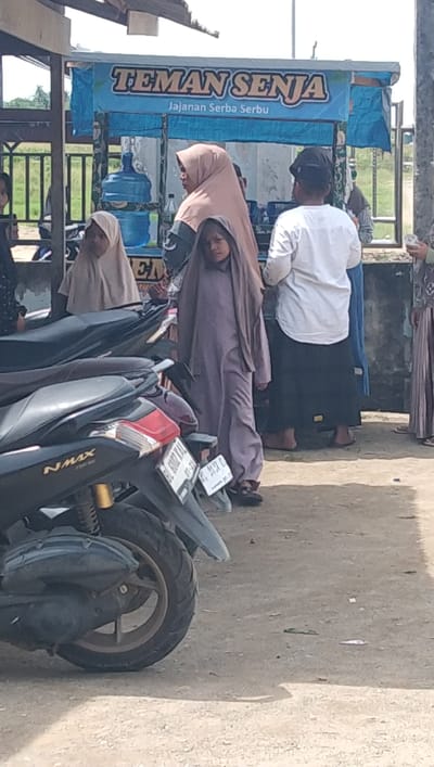 The image depicts an outdoor scene in Dayah Baroh, Indonesia, featuring several people and motorcycles. In the foreground, two parked motorcycles are visible, one black with the "NMAX" logo clearly displayed. Behind the motorcycles, a group of individuals is gathered near a stall. A young girl in a purple hijab stands prominently, looking towards the camera. To her left, a woman in a light brown hijab is partially visible, and another child in a similar hijab is behind her. To the right of the young girl, a man wearing a white shirt, black cap, and a sarong stands facing away from the camera. Further to the right, another person in a patterned outfit is partially visible. The stall behind them has a blue banner with text that appears to read "TEMAN SENJA" and "Jajanan Serba Serbu." A large blue water dispenser is also visible behind the stall. The ground is dirt and gravel, and the overall setting appears to be a roadside or market area.