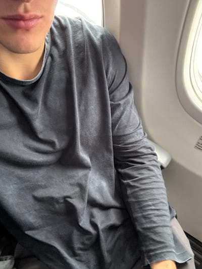 The image shows a person sitting in an airplane seat. The person is wearing a dark gray long-sleeved shirt. The person's lips and part of the neck are visible. The person's arm is resting on the window ledge and is wrinkled. The airplane window can be seen in the upper left corner of the image, along with a portion of the seat.