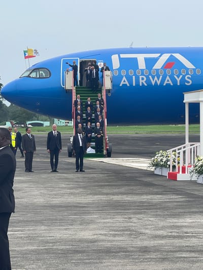 The image captures a formal arrival or departure ceremony on an airport tarmac. A large, bright blue airplane, clearly branded "ITA AIRWAYS" in white lettering on its fuselage, dominates the left side of the frame. The plane appears to be an Airbus model, with its nose cone and cockpit visible on the far left.

A mobile airstair, adorned with elegant white and green floral arrangements and subtle red accents, is positioned at the main passenger door of the aircraft. On these stairs, a group of individuals in dark suits and official uniforms, including pilots and flight attendants, are visible, some seemingly interacting with a person at the top of the stairs. Below, on the tarmac, several men in dark suits stand in a structured line, extending from the base of the stairs to the foreground.

Near the cockpit window, two flags are proudly displayed: the yellow and white flag of Vatican City with its papal insignia, and another flag with yellow, blue, and red horizontal stripes. In the middle ground, to the right of the plane, a white structure, also decorated with flowers, suggests a ceremonial path or red carpet setup. The ground is a grey tarmac, and the sky is overcast, indicating daytime. The scene suggests a significant event, likely an official visit or diplomatic mission, possibly involving a high-ranking Vatican official, taking place at an airport in Malabo, Equatorial Guinea. The visible text on the plane is "ITA AIRWAYS".