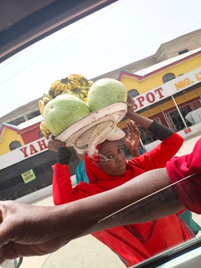 The image, captured from inside a vehicle in Hotoro, Nigeria, depicts a dark-skinned woman balancing a worn white tray on her head. The tray contains two green watermelons and a large cluster of yellow bananas. The woman wears a red hooded garment and a nose stud, looking directly at the camera with her hands raised, appearing to stabilize the load. Behind her stands a yellow building with red trim, displaying partial red lettering "YAHYA SPOT NIG. LTD" and a green "NOW OPEN" sign. A second individual in a blue shirt is partially visible behind the woman. A dark-skinned arm is present in the lower left foreground, extending from the camera's perspective. The scene is illuminated by bright daylight.