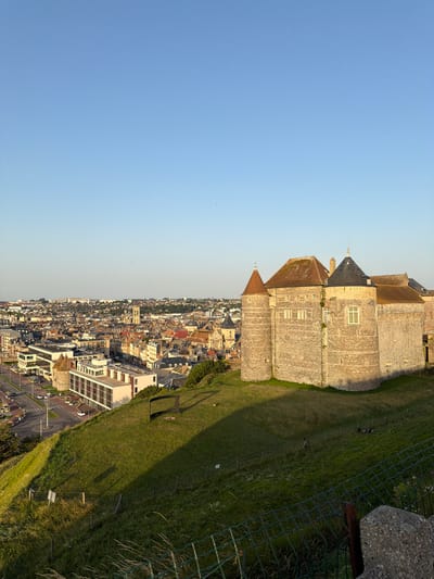 The image depicts a panoramic view of Dieppe, France, taken from a high vantage point. The scene is divided diagonally, with a vast expanse of clear blue sky occupying the left half and the town and its surroundings filling the right. Dominating the upper right portion of the frame is a large, stone castle with multiple turrets and pitched roofs, suggesting historical significance. The castle is perched on a grassy hillside, which slopes down towards the densely packed urban landscape.
The town below is a mosaic of buildings with varied architectural styles, featuring terracotta roofs and light-colored facades. A modern, multi-story building with large windows is prominently visible in the foreground of the town, contrasting with the older structures. Roads and a roundabout are visible, with a few cars suggesting some level of activity, though the overall impression is one of a quiet afternoon. The setting appears to be a coastal town, given the likely location of Dieppe.
The lighting indicates that it is either late afternoon or early evening, with long shadows cast across the grassy slope, and the sky showing a gentle gradient of color. A wire fence runs along the edge of the grassy area, partially obscuring the view of the town and the grassy slope itself. The overall mood is serene and picturesque, offering a comprehensive look at the town and its historical landmarks. There is no visible text in the image.