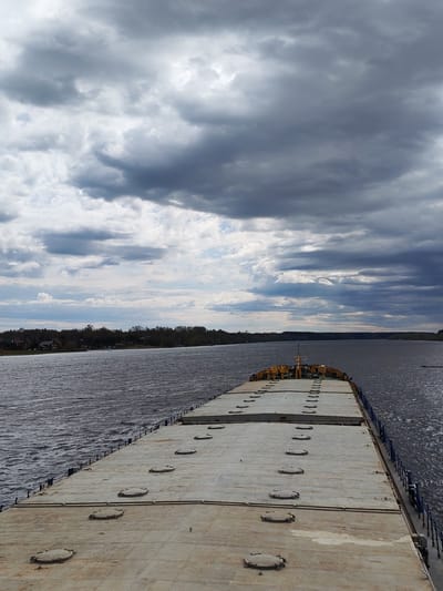 The image depicts the deck of a barge or similar large flat-topped vessel, with a series of round access hatches visible on its surface. The vessel is traversing a wide body of water, presumably a river or large lake. The shoreline is visible in the distance, with trees and some structures that might be houses. The sky is overcast with heavy, dark clouds. The overall impression is one of a cloudy day on a large body of water. The location is Mikheevo, Russia.