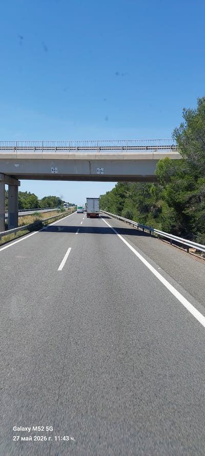The image depicts a highway in Vandellòs i l'Hospitalet de l'Infant, Spain. The camera is oriented to the right, capturing a view of the road stretching forward. A concrete overpass with posted clearance heights of 5.06m and 5.15m spans the highway. Vehicles, including a truck, are visible in the distance on the road. The sky is clear and blue on the left side of the frame, while trees line the right side. The road surface is asphalt with white lane markings. A metal guardrail runs along the edge of the highway. Text from the camera is visible at the bottom of the image indicating "Galaxy M52 5G, 27 mai 2026 r. 11:43".