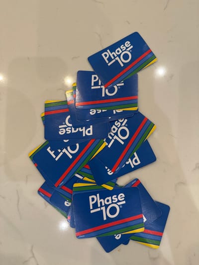A collection of approximately 20-30 "Phase 10" playing cards is scattered and overlapping on a light-colored, smooth surface, possibly a countertop, in the city of McAllen, United States of America. The cards are uniformly blue on their backs, each displaying the white "Phase 10" logo, consisting of the word "Phase" above a horizontal line and "10" below it. Four horizontal colored stripes—red, blue, green, and yellow—are present near the bottom edge of each card's back. The cards are arranged haphazardly, with various orientations, and some partially overlap others. No individuals, actions, or interactions are present in the image. The surface exhibits a subtle veined pattern consistent with natural stone or laminate. Illumination appears to be from an overhead light source, creating a bright and even exposure across the scene.