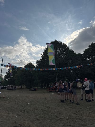 A daytime outdoor scene shows people standing on a grassy field under a partly cloudy sky. Tall trees form a backdrop. Colorful flags and banners are strung between poles, adding a festive atmosphere. The people are a mix of adults and what look like some teenagers. The sky has a patch of puffy grey clouds near the tree line.