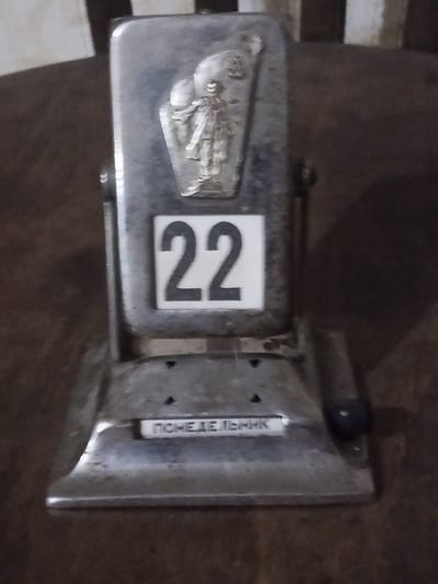 An antique desktop perpetual calendar is displayed on a dark wooden surface. The calendar has a metallic base and a rotating upper section that shows the number "22" in black digits on a white background. Above the number "22," there is a relief image of a person in a tunic holding a staff or torch. The base of the calendar has a metallic plaque with text in Cyrillic script that reads "ПОНЕДЕЛЬНИК," meaning "Monday." The background is blurred, but appears to be an indoor setting with vertical lines, possibly representing blinds or textured wallpaper. The image's lighting is dim, with reflections visible on the metallic surfaces of the calendar. The calendar appears to be an artifact of historical or sentimental value. There is no information within the image to indicate its specific location in the city of Dubossary, Moldova.