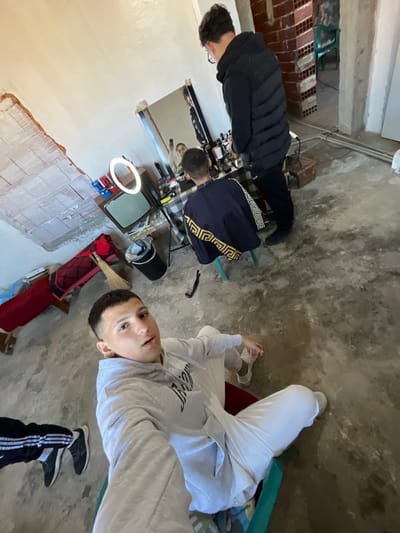 The image depicts an indoor scene in the city of Hvostiane, Bulgaria. In the foreground, a young male with short, dark, faded hair, wearing a light grey hooded sweatshirt and white pants, is seated and taking a selfie, looking directly at the camera. To his right, a male client is seated on a green stool, facing away from the camera and towards a rectangular mirror. The client wears a dark blue barber cape with a gold Greek key pattern along its trim. Behind the client, a male wearing a dark puffer vest and glasses stands, his reflection in the mirror shows him holding a mobile phone.

The mirror is positioned on a table covered with a plaid cloth, displaying various bottles, containers, and an illuminated ring light. To the left of this setup, an older grey cathode-ray tube television sits on a low surface, adjacent to a black cylindrical trash can with a silver lid. A traditional broom and some folded red fabric are on the concrete floor to the left. A black, U-shaped object, possibly a hair clipper or comb, rests on the concrete floor between the foreground subject and the client.

The room features a textured concrete floor and unfinished walls; the wall on the left exhibits exposed plaster or concrete with visible markings, while a doorway on the right reveals exposed red brickwork and a green chair in an adjacent area.