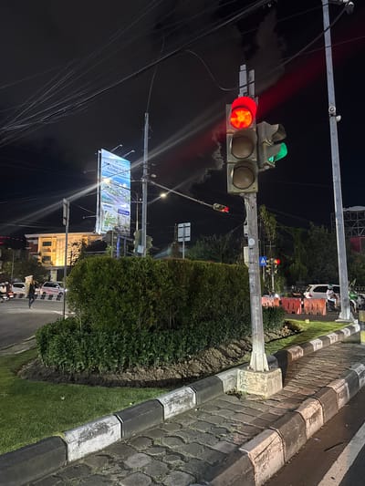 The image depicts a street scene at night in Denpasar Barat, Indonesia. A traffic light is prominently featured, with its red light illuminated, indicating a stop. The traffic light is mounted on a metal pole, which is part of a larger street infrastructure. In the background, a billboard with the text "MORE THAN A HOME, A LIVING ASSET" is visible, advertising a real estate development. The billboard features images of a modern building and a tranquil landscape.
The scene includes a paved road with a distinct curb made of stone blocks, interspersed with black and white sections. A patch of green grass runs alongside the road, bordered by dense green bushes. In the distance, trees and some buildings with lights on can be seen, suggesting an urban or suburban environment. A few vehicles, including what appears to be a white car and a motorcycle, are present on the road, indicating ongoing traffic. The sky is dark, consistent with nighttime. The overall impression is of a typical urban setting at night, with the traffic light and billboard being the most striking elements. There is no indication of specific weather conditions. The atmosphere appears functional and everyday.