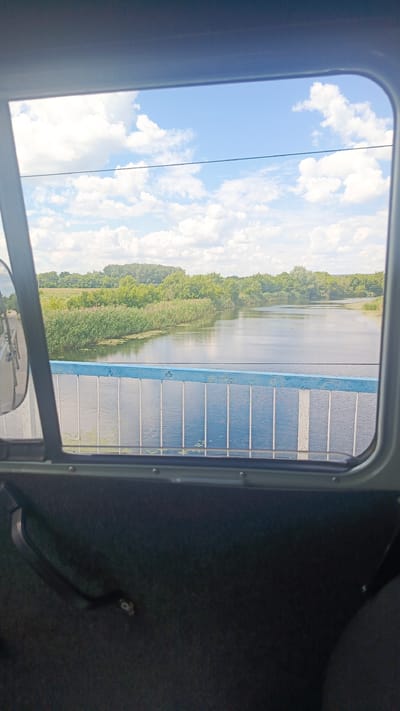 The image, taken from inside a vehicle through a window, shows a view of a river on a cloudy day. A bridge with a blue and white railing spans the river. The riverbanks are lined with lush green trees and reeds. The sky is filled with white, fluffy clouds against a bright blue backdrop. The interior of the vehicle, including part of a dark-colored door panel and a handle, is visible in the foreground. The location is the city of Rybushka, Russia.