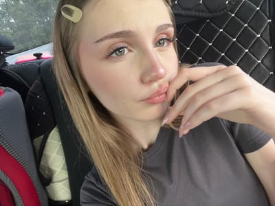 A young woman with blonde hair and a light skin tone is taking a selfie in a car. She is wearing a grey t-shirt and has on makeup, including mascara and blush. Her right hand is touching her lips. The background of the image shows the interior of the car, including a black quilted seat cover and a portion of a seat with red and white fabric. The overall environment appears to be an indoor setting, possibly a vehicle in a suburban or rural area, given the glimpse of trees visible through the car window. There is no indication that this scene is located in Bairak, Russia.