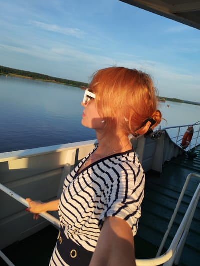 A woman with red hair is standing on the deck of a boat, looking out at a body of water. She is wearing a striped shirt and white sunglasses. The sun is setting, casting a warm glow on the scene. The water is calm, and the sky is a clear blue with a few wispy clouds. The boat appears to be in a rural area, with trees visible on the distant shore.