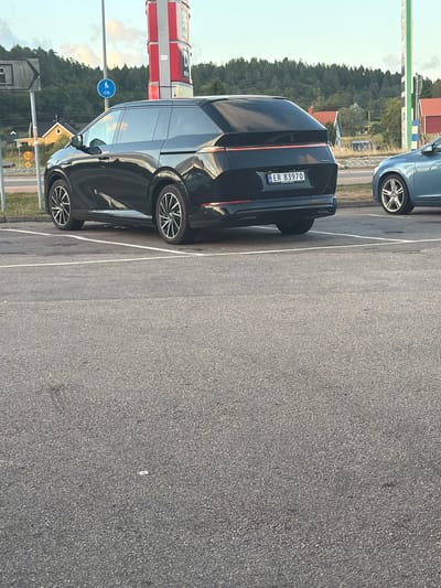 In Hogstorp, Sweden, a black electric car is parked in a lot. The car's license plate reads "ER 83970". A blue car is parked to the right of the black car. In the background, there are trees, houses, and a tall red and white structure that appears to be a gas station sign. A speed limit sign indicating 50 is visible on the left.