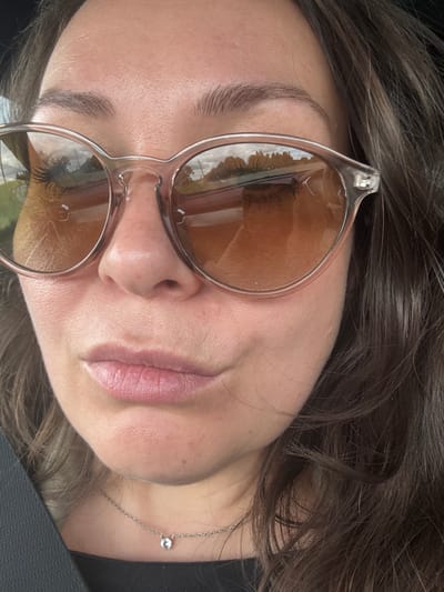 A close-up, waist-up, outdoor selfie of a woman wearing sunglasses and a necklace, looking into the camera. The woman has dark brown hair, fair skin, and is wearing a black top. She is wearing light pink lipstick and her eyebrows are dark brown and arched. The sunglasses have clear frames and amber lenses. The background is blurred, showing trees and a road. The image is likely taken in Krasnoyarsk, Russia, given the reference to the city in the prompt.