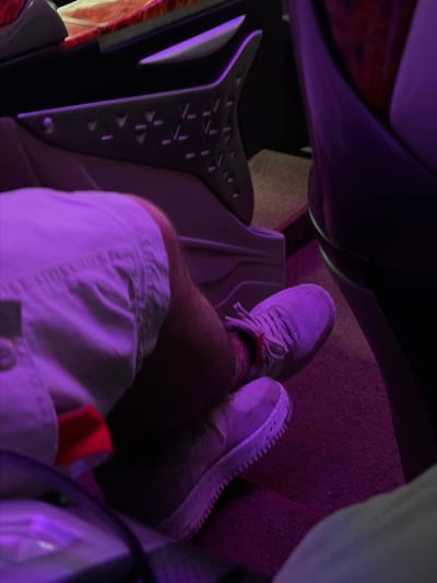 The image shows a person's legs and feet inside what appears to be a vehicle's cabin, possibly an airplane or a luxury car. The interior is illuminated with purple lighting, creating a distinct ambiance. The person is wearing light-colored shorts and light-colored athletic shoes with laces. The shoes have a textured sole. In the background, there is a dark-colored seat and part of the vehicle's interior paneling, which has a patterned design and some metallic accents. The flooring appears to be carpeted in a reddish-purple hue. There are no visible signs of the city of Thôn 6, Vietnam in the image, as the setting is entirely enclosed within a vehicle.