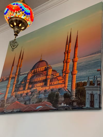 The image shows a large canvas print of the Blue Mosque in Istanbul, Turkey, hanging on a white wall. A colorful mosaic Turkish lamp hangs from the ceiling in the upper left corner of the frame, partially obscuring the top left corner of the canvas. The canvas print depicts the Blue Mosque at sunset, with its domes and minarets illuminated by the warm, golden light. The sea is visible in the background. The print is mounted on a white wall, with a white ceiling visible at the top of the frame. The scene appears to be an interior of a room, possibly a home or a business, in Akron, United States of America. There are no people visible in the image.