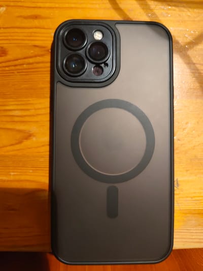 The image shows the back of a black iPhone case, lying on a wooden surface. The case has a camera bump with three lenses in the upper left corner. In the center of the case, there is a circular magnetic charging ring, and below that, a small rectangular magnetic alignment indicator. The wooden surface appears to be a table with visible grain and some slight wear. The lighting suggests an indoor environment, possibly a home or office, and there are no people or other objects visible in the frame. The location is not specified in the image.