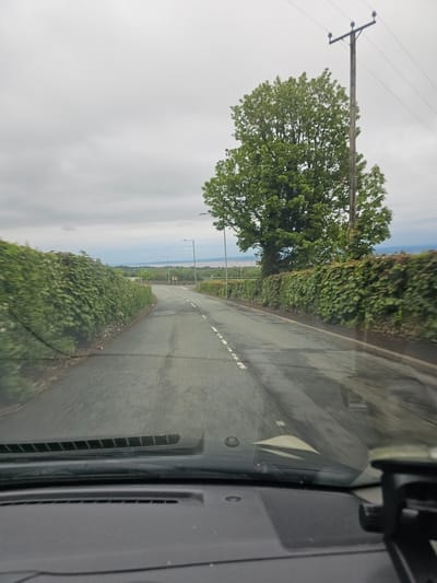 The image depicts a view from inside a vehicle, likely a car, traveling on a road. The road is asphalt and has a center line of dashed white markings. The road is lined with green hedges on both sides. A large tree is visible on the left side of the road, with a utility pole located nearby. In the distance, the sea and horizon are visible under a cloudy sky. The location is likely in Halkyn, United Kingdom.