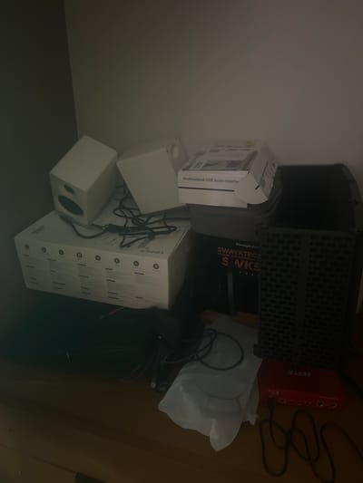 The image shows a cluttered shelf with various electronic equipment and packaging. In the center of the image, two white computer speakers are stacked on top of a white cardboard box. To the right of the speakers, a black, rectangular device with a textured surface is visible. In front of the black device and speakers, there are several black cables and a white plastic bag. A red audio interface with ports and buttons is positioned in the lower right corner of the image, with black cables connected to it. The background is a plain, light-colored wall. The scene is illuminated by a soft light, creating shadows on the shelf and surrounding objects. There are no people visible in the image, and no specific location in the city of Mussolini, Italy can be determined from the provided visual information.