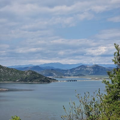 The image depicts a body of water (likely Skadar Lake) extending into the distance, flanked by mountains on the left and greenery in the foreground on the right. A causeway with a bridge segment stretches across the lake. The sky is partly cloudy with a blue hue. The location is likely Godinje, Montenegro, given the landscape characteristics.