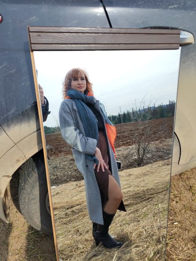 A woman is reflected in a mirror propped against a dark-colored vehicle. She is wearing a grey coat, a dark blue scarf, and black boots. She has reddish-brown hair. The mirror reflects a rural landscape with a field of dry grass and a plowed field in the background, along with some trees and power lines. The location is Нечкино, Russia. A portion of the vehicle, including a tire, is visible.