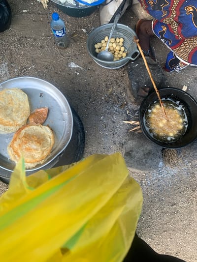 This is a high-angle, medium shot of an outdoor food preparation scene in Reak, Nigeria. The ground is composed of dirt and gravel, with scattered stones and dry vegetation. In the foreground, a bright yellow plastic bag obscures the lower portion of the frame, with diagonal green stripes visible.

To the left, a metal tray holds several flat, golden-brown fried food items, possibly pancakes or fritters, and a small portion of a reddish paste. Adjacent to the tray is a black tire, serving as a base.

In the center of the image, a metal colander is filled with small, spherical, light-colored items, resembling chickpeas or small dumplings. A metal ladle rests inside the colander. A clear plastic bottle containing a brownish liquid and a blue cap sits on the ground nearby, with a single piece of straw extending from it.

To the right, a dark pot is placed on what appears to be a makeshift stove with rocks. The pot contains bubbling hot oil with small, golden-brown food items being fried. A long, thin wooden stick is immersed in the oil, stirring the frying food. Partially visible at the top right is the leg of a person wearing a colorful, patterned fabric with a blue, red, and yellow design. The scene suggests a casual, outdoor cooking environment.
