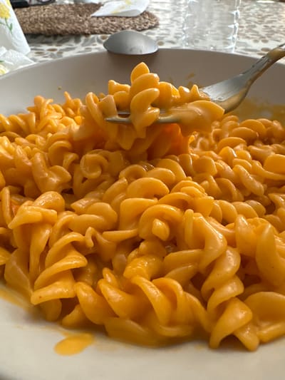 The image shows a close-up view of a bowl of rotini pasta with a cheesy orange sauce. A fork is lifting a portion of the pasta from the bowl. The pasta is coated in a smooth, creamy sauce that appears to be cheese-based, giving it a vibrant orange hue. The background is slightly blurred, showing a placemat, a napkin, a spoon, and a plastic water bottle, suggesting a casual dining setting. There are no discernible landmarks to indicate a specific location within Macclesfield, United Kingdom.