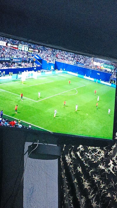 The image captures a televised soccer match displayed on a screen. The screen shows a game between two teams, indicated by the score graphic that reads "ARG 0 1 EGY". The field is green, with white lines marking the boundaries and penalty areas. Several players in red and white uniforms are visible on the field, engaged in the game. Spectators are present in the stands, filling the stadium. The broadcast also includes overlay information such as "WORLD CUP 2026" and "SPORTS". The screen is mounted on a wall, with a dark textured surface visible in the foreground, suggesting the image was taken in a room. The location within "Пистамазор, Tajikistan" is not discernible from the image itself.