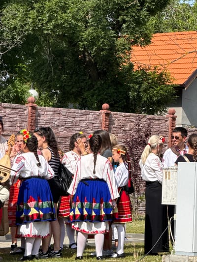 In the city of Pordim, Bulgaria, a group of people are gathered outdoors, dressed in traditional Bulgarian folk costumes. The costumes feature white embroidered blouses and colorful skirts with geometric patterns. Some individuals are wearing flower headbands. A person on the left is holding a stringed instrument, possibly a kaval or gudulka. The background includes a stone wall, lush green trees, and a house with a red tiled roof. The atmosphere appears festive, suggesting a cultural event or celebration.