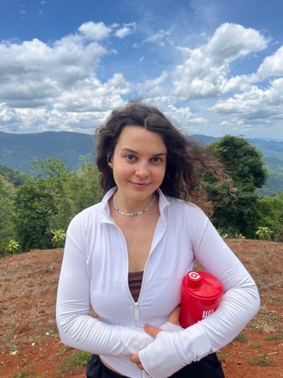 A woman with curly brown hair stands outdoors holding a red water bottle. She is wearing a white zip-up jacket over a brown top and black shorts. She has on a beaded necklace. Behind her is a mountainous landscape with green trees and a blue sky with white clouds. The ground in front of her is reddish-brown dirt with sparse vegetation.