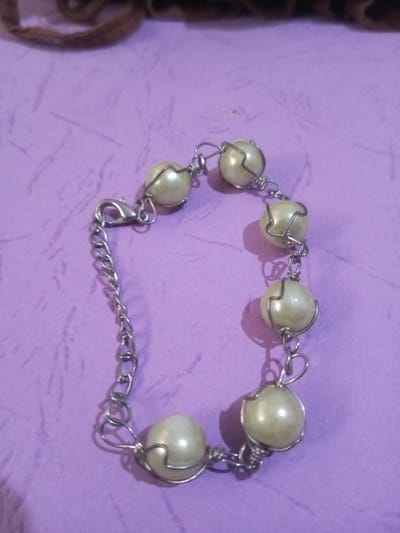 A single piece of jewelry, identified as a bracelet, is displayed on a textured, light purple surface. The bracelet consists of nine spherical, off-white beads, each individually encased in thin, silver-toned wire loops and interconnected to form a chain. One end of the bracelet features an additional segment of silver-toned metal chain, terminating in a lobster claw clasp and an extender chain. A blurred, dark brown object is partially visible in the upper background. The scene is located in Chandragiri, Nepal.