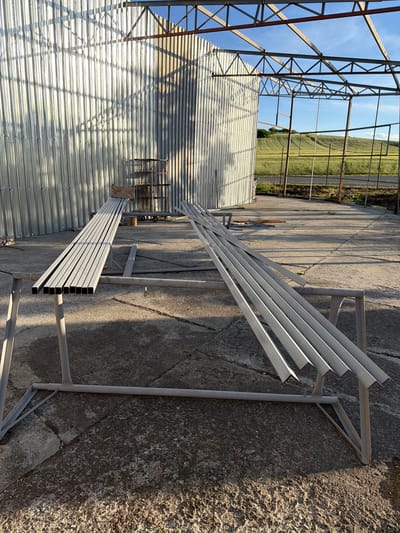 The image depicts a partially constructed metal structure, likely a greenhouse or agricultural building, in what appears to be a rural or semi-rural location. The frame of the structure is visible, constructed of metal beams and supports, with corrugated metal siding partially installed on one side. Metal pipes are placed on the side of the building. The ground is a paved surface with cracks and wear. In the background, a field or grassy area can be seen, suggesting the surrounding environment is agricultural. The location is Dabnik, Bulgaria. There are no people visible in the image.