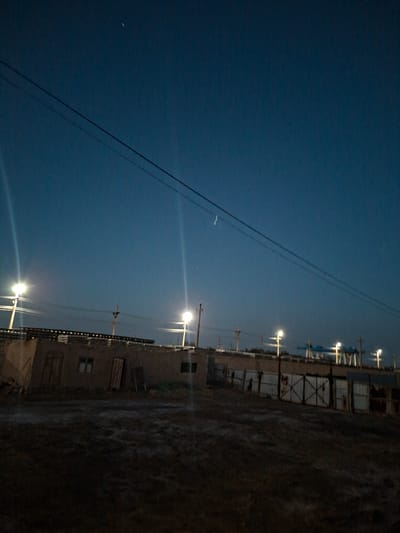 The image depicts an outdoor scene at dusk or night in Kungrad, Uzbekistan. The sky is a deep blue, with power lines crisscrossing horizontally across the upper portion of the frame. Several streetlights are illuminated, casting bright beams of light. In the foreground and mid-ground, there are low-lying, weathered structures that appear to be buildings or sheds, constructed of brick or mud-brick with visible doors and windows. A dirt or gravel area is in front of these structures. In the background, further industrial-looking structures and more streetlights are visible, along with some distant, illuminated cranes. The overall impression is one of a rural or semi-industrial area at twilight. No people are visible in the image.