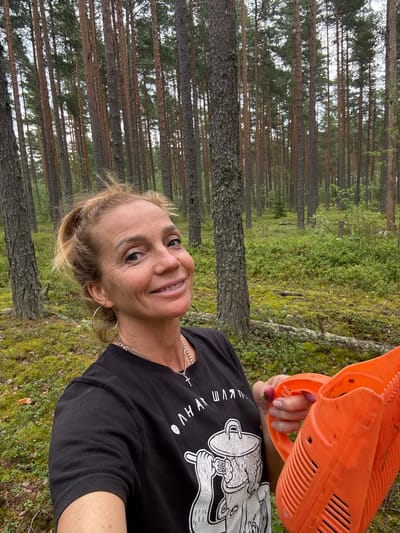In Kliuchevoe, Russia, a middle-aged Caucasian woman with blonde hair pulled back in a ponytail and a light tan is shown in a forest. She is wearing a black t-shirt with a graphic print of a character holding a teapot and a silver necklace with a cross pendant. She is also wearing large hoop earrings. The woman is smiling and holding an orange berry-picking tool in her right hand. The background is a dense pine forest with a carpet of green moss and low-lying shrubs covering the ground. The lighting suggests it is daytime, with sunlight filtering through the trees.