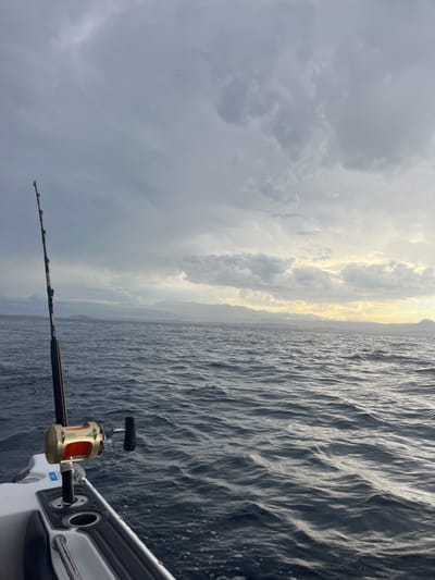 The image depicts a fishing rod extending from a boat towards the sea. The ocean, covered in ripples, stretches toward a distant shoreline under a sky filled with dark, brooding clouds. A sliver of sunlight breaks through the clouds, casting a faint glow on the water and the distant land. The fishing rod, held firmly in a boat-mounted holder, suggests anticipation. The overall atmosphere is one of maritime activity under overcast conditions.