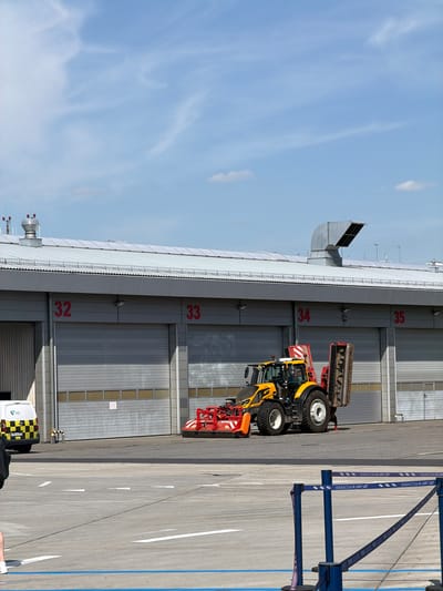 The image shows an outdoor scene at an airport, likely in Balice, Poland, given the text "KRAKÓW AIRPORT" visible on blue and purple banners. The day appears to be bright and sunny with a clear blue sky and scattered white clouds.
The main subjects are a yellow and red industrial tractor with a large, rotating brush attachment parked in front of a row of large, grey hangar doors. The doors are numbered with red digits: 32, 33, 34, and 35 are clearly visible. The tractor is a significant object in the frame, positioned to the left of the center.
In the foreground on the right, a blue structure with purple banners stretches into the distance, indicating a boundary or pathway. The ground is paved, with white markings and blue lines visible, suggesting it's an airport apron or taxiway. A white van with a black and yellow checkered pattern on its rear is parked to the left, near the hangar doors.
No people are clearly visible in the image, but their presence is implied by the equipment and the overall setting. The scene appears to be a functional area of the airport, possibly where maintenance or ground operations take place. The overall atmosphere is one of a clear, operational day at an international airport.