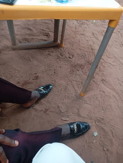 The scene depicts an overhead view of a ground area under a table, situated on reddish-brown compacted dirt, possibly an outdoor or unpaved indoor setting in Okpatu-Ukehe, Nigeria. Visible on the dirt are indistinct footprints and scattered small fragments of debris. A table with a yellow-colored edge is partially present in the upper frame. Its legs are gray metal; one leg features a yellow plastic cap at its base, while another is connected to a beige-colored lower crossbar. On the visible portion of the tabletop, a dark rectangular object and a blue plastic cap are present. Two individuals are seated, with only their lower legs and feet visible. One individual, positioned on the left, wears dark maroon trousers and a black, patent leather-like loafer with a gold-colored chain embellishment. A dark-skinned hand rests on this individual's left thigh. The second individual, positioned in the lower right, wears off-white trousers and a matching black, patent leather-like loafer with a gold-colored chain embellishment. The exact location within Okpatu-Ukehe is not discernible from the provided image.