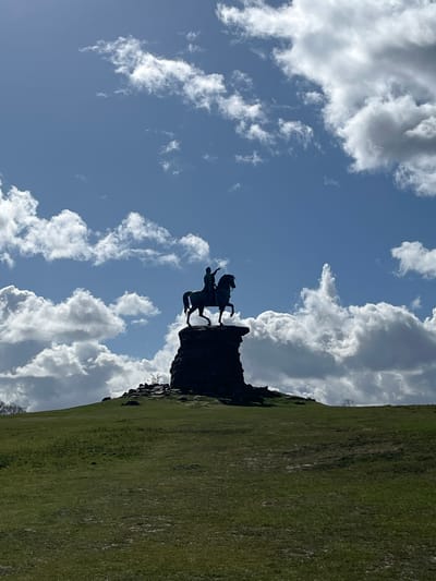 The image presents a silhouetted equestrian statue situated atop a grassy hill under a blue sky populated with white cumulus clouds, located in The Village, United Kingdom. The central subject is an imposing statue, appearing dark against the bright sky, depicting a figure on horseback. The horse is captured in a rearing pose on its hind legs, while the rider, also silhouetted, has one arm raised and extended, pointing forward and upward. This statue is mounted on a substantial, dark, rocky pedestal that integrates with the summit of the verdant hill. The foreground consists of a gently sloping expanse of green grass with some lighter patches. The sky, occupying the upper two-thirds of the frame, displays varying cloud formations, some brightly lit and others casting subtle shadows, with the sun appearing to be behind the statue, creating the pronounced silhouette effect. No people or discernible structures beyond the statue and its base are visible.