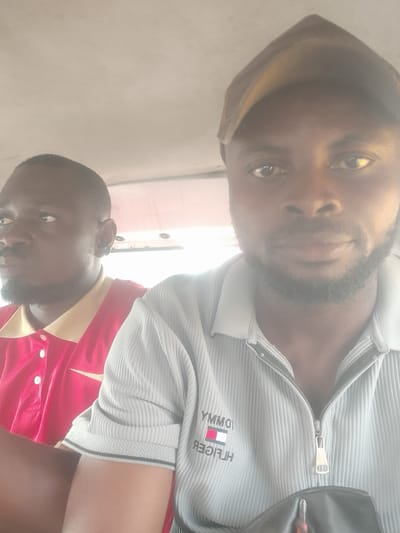 Two Black men are in the foreground of the image, seemingly inside a vehicle. The man on the right, wearing a brown cap and a light gray ribbed shirt with a zipper and a Tommy Hilfiger logo, is looking directly at the camera. The man on the left, wearing a red collared shirt, is looking to the left. Both men have dark skin and beards. The environment appears to be the interior of a car or bus, with a light-colored ceiling visible. The lighting is bright, suggesting it is daytime. The image is slightly blurred, particularly around the edges. There is no specific indicator within the image itself to confirm the location as Alagbede, Nigeria.