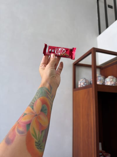 A person's tattooed arm is extended towards the viewer, holding a red Kit Kat candy bar. The tattoo features a floral design with shades of orange, pink, and green, with hints of purple. The Kit Kat wrapper is red with the iconic white "Kit Kat" logo. The background is a bright, plain wall, with a wooden shelf unit visible in the upper right portion of the image, containing decorative ceramic cups. The overall impression is casual and domestic. The lighting suggests daytime, possibly indoors. The scene is static, with no active event occurring other than the presentation of the candy bar. There is text on the Kit Kat wrapper, but it is not legible in detail. However, the "Nestle" logo is visible.
