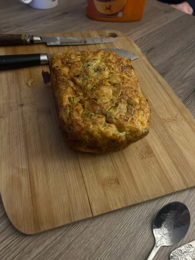 The image presents a close-up, overhead view of a savory loaf of food resting on a wooden cutting board. The loaf is golden brown with a textured, somewhat craggy surface, showing flecks of green, possibly herbs or chopped vegetables, embedded within. Its shape is roughly rectangular or oblong, consistent with a quick bread or cake baked in a loaf pan.
To the left of the loaf, on the cutting board, are two knives. One has a serrated blade and a handle with visible grain, possibly wood or a similar material, in shades of brown. The second knife, partially obscured by the first, has a black handle. Both knives appear ready for use, suggesting the loaf is either about to be sliced or has just been removed from the oven.
The cutting board itself is made of light-colored wood, likely bamboo, with distinct parallel lines of wood grain. It shows some faint marks and cuts on its surface, indicating previous use.
In the upper right corner of the frame, two silver-colored utensils are partially visible, resting on a gray surface next to the cutting board. They appear to be spoons or serving utensils, distinguished by ornate, embossed patterns on their handles and bowls.
In the upper left portion of the image, an orange-colored bottle or container is partially visible, with a label that is too blurry to read. Below it, in the bottom left corner, a small portion of a white object, possibly a cup or ceramic dish, can be seen.
The background consists of a gray surface with a subtle wood-like grain pattern, suggesting a countertop or floor. The overall setting appears to be a casual indoor environment, likely a kitchen, where food preparation or serving is taking place.