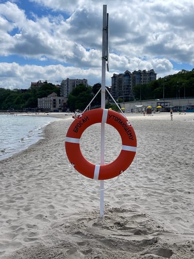 The image depicts a beach scene in Pionersky, Russia, under a partly cloudy sky. The main subject in the foreground is an orange lifebuoy with two white stripes, mounted on a white pole. The lifebuoy features Cyrillic text: "УТОПАЮЩЕМУ" (To the drowning one) inscribed along its top curve and "БРОСАЙ" (Throw) along its bottom curve, indicating its use for rescue.
Beyond the lifebuoy, a wide stretch of light-colored sand extends towards calm water with gentle waves. The sand shows rippled patterns and some footprints. In the midground, several people are visible on the beach, enjoying the day, though their specific activities are indistinct. Yellow beach umbrellas are scattered across the sand further down the shore.
The background features a line of multi-story buildings, likely hotels or residential complexes, peeking through a dense cover of green trees. One of the cropped views reveals a construction crane in the distance among these buildings. The sky is bright with patches of blue and large, cumulus clouds. The scene suggests a pleasant, active daytime setting at a coastal resort or public beach.
