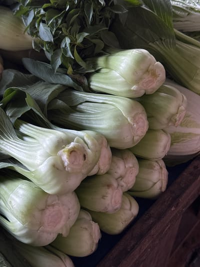 A close-up, overhead shot showcases a pile of fresh bok choy, a type of Chinese cabbage, bundled together and resting in a wooden crate. The bok choy is characterized by its thick, white stems and vibrant green leaves, with some showing slight bruising or imperfections. The scene is lit from above, casting shadows that highlight the texture and shape of the vegetables. The background is dark and out of focus, with hints of other produce, possibly more leafy greens. This image appears to be taken at a market stall in the city of Karangsambung, Indonesia, where fresh produce is typically displayed for sale.