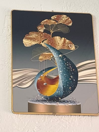 The image shows a framed piece of abstract art hanging on a textured white wall. The artwork features a dark background with a gradient transitioning to a lighter gray at the bottom. In the upper portion of the artwork, there are several stylized golden-brown leaves with small, sparkling embellishments. Below the leaves, a prominent, tear-drop shaped object dominates the center of the frame, colored in shades of blue, yellow, and red. This object is adorned with more sparkling embellishments. The base of this central element rests on a gold-colored platform, which itself is sitting on a gray surface. Flowing lines of white and beige fabric or ribbon are depicted in the background, creating a sense of movement. The artwork is enclosed in a thin, gold-colored frame. The location of this artwork is not provided in the image or its context.