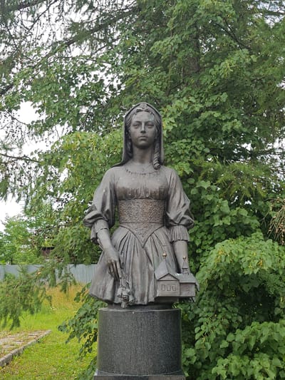 This is a medium shot, eye-level photograph of a bronze statue of a woman in front of lush green trees. The statue depicts a woman in historical attire, wearing a long dress with puffed sleeves and an ornate, patterned bodice. She has a headpiece or veil. In her left hand, she holds a bouquet of roses, and in her right hand, she holds a miniature model of a building with a peaked roof and windows. The statue stands on a dark, granite pedestal. The background consists of dense green foliage, suggesting an outdoor setting, likely a park or garden. There is a glimpse of a gray corrugated metal fence and some dry grass to the left of the statue, indicating it's an outdoor public space. The sky is overcast, with a white, hazy appearance. The statue appears to be located in the city of Ulitkino, Russia, as suggested by the context.