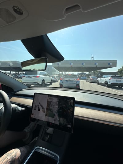 A first-person perspective captures the interior of a Tesla vehicle and the view through its windshield, depicting a queue of vehicles at a multi-lane international border crossing under bright, sunny conditions. The location is identified as Kapitan Andreevo, Bulgaria.
The observer is positioned in the driver's seat of a Tesla, indicated by the steering wheel, wooden dashboard trim, and a prominent central touchscreen display. A tattooed left forearm and hand are visible on the lower left, along with parts of the driver's legs wearing light-colored sneakers, resting on the pedals. The touchscreen displays a map interface with traffic conditions, showing several vehicle icons, and a destination of "22 km." Music playback for "Elton John" is also visible on the screen. An overhead console with a speaker grille and a small sensor/camera is seen above the rearview mirror.
Through the windshield, a line of cars is visible ahead, stationary or moving slowly in multiple lanes leading to border control booths. Directly in the rearview mirror, a white Mercedes-Benz GLS SUV with German license plate "M RQ 580" is visible. In the lanes ahead, a grey Subaru XV/Crosstrek with Bulgarian license plate "CB 4213 XM" occupies the left lane, while a white SUV (possibly a Voyah Free) with Bulgarian license plate "PB 2293 TE" is in the right lane. A dark sedan is partially visible behind the Subaru.
Overhead, a large gantry spans the width of the road, marked with distinct signage for different vehicle types and border procedures. Text on the gantries includes "CARS," "BUSES," and "КАМИОНИ" (Cyrillic for "TRUCKS"), along with other Cyrillic script. Green lights are visible on some of the gantry structures, indicating open lanes. The overall infrastructure suggests a busy and established border crossing. The sky is clear blue with no clouds. Various buildings associated with the border facility are visible in the background, including one with a small antenna tower on its roof.