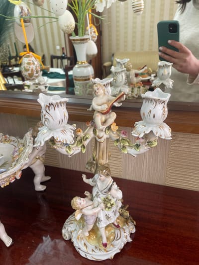 The image depicts a porcelain candelabra resting on a dark brown wooden surface. The candelabra features a central figure of cherubic forms amidst foliage and floral candle holders. The reflection in a nearby mirror shows a hand holding a green smartphone taking the picture, Easter eggs, a decorative vase, and a glimpse of the room's interior. The exact location within the city of Akhalkalaki, Georgia, cannot be determined from the provided image.