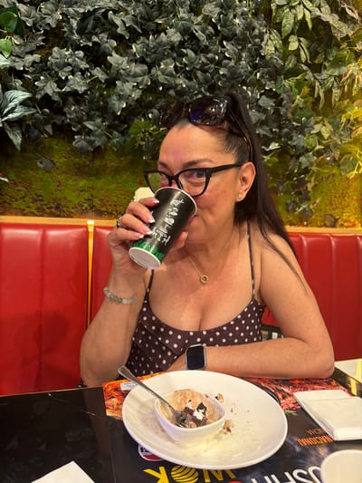 A woman with long dark hair pulled back and wearing glasses is sitting at a table with a red booth behind her and a wall of artificial plants. She is drinking from a black and green cup with white polka dots. On the table in front of her is a white plate with a partially eaten dessert and a spoon. She is wearing a polka dot dress and a gold necklace. There is a smartwatch on her left wrist and a bracelet on her right wrist. The location is specified as Carretera De San Martín, Spain.