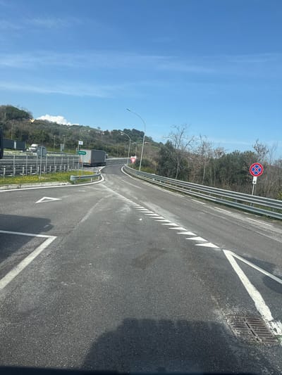 The image presents a daylight scene of a road junction in Tortoreto, Italy, under a mostly clear blue sky with sparse clouds. The foreground features an asphalt roadway with distinct white road markings, including a solid line on the left, a series of white triangular yield markings extending towards the center, and a white V-shaped marking bordering a metal drainage grate on the right.

To the left, a grassy median strip with small yellow flowers separates a primary lane from an intersecting roadway. Two dark-colored tractor-trailers are visible in the mid-ground. One truck, predominantly dark grey with a light grey rear door, is positioned on a curved connecting road, appearing to navigate a left turn. Adjacent to this truck, a blue rectangular sign displays "ANCONA" in white text with a right-pointing arrow. Another dark truck is partially visible on the far left behind a metal guardrail.

To the right, the road gently curves uphill, bordered by a multi-segment metal guardrail. A red triangular "GIVE WAY" sign is affixed on a pole above the guardrail. Further down the road on the right, a red circular "NO STOPPING" sign with a downward-pointing arrow is visible. A solitary streetlight pole extends upwards from the right side.

The background consists of undulating hills covered with a mix of bare deciduous trees and evergreen foliage. Faint structures are discernible on the distant hillsides. The bright sunlight casts shadows from the viewing vehicle onto the foreground asphalt, indicating a mid-day lighting condition. No persons are present in the scene.
